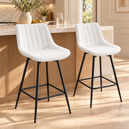 Deux chaises hautes de bar blanches à dossier rembourré et surpiqûres verticales, avec pieds noirs, devant un îlot en bois et pierre dans une cuisine chaleureuse.