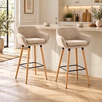 Lot de deux tabourets de bar scandinaves beiges avec accoudoirs et pieds en bois devant un comptoir de cuisine crème, dans un intérieur chaleureux et lumineux.